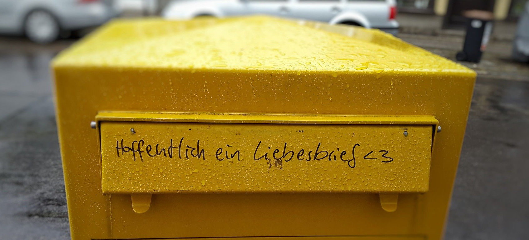 Eine Fotografie die einen alten gelben Briefkasten zeigt. Der Briefkasten steht für Kontakt - er ist regennass und auf dem Deckel über dem einwurfschlitz steht mit schwarzen Edding stift geschrieben: "Hoffentlich ein Liebesbrief!"