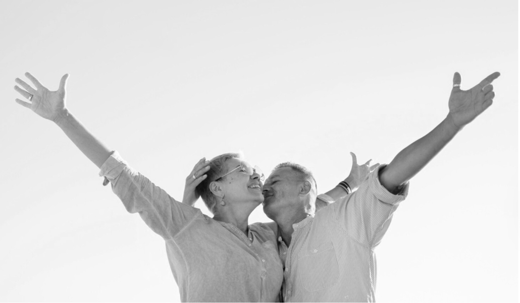 Schwarzweiss Fotografie von einem Paar welches sich umarmt und mit dem jeweils frein Arm nach oben zeigt. Beide machen einen glücklichen Eindruck. Es steht für das Win-Win Ergebnis einer gelungenen Mediation